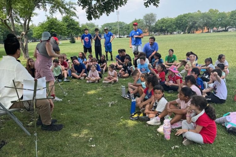 Children sit on grass in a park listening to two adults.